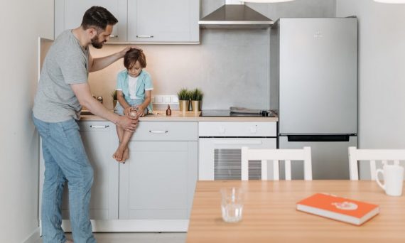 A father assists his son sitting on a kitchen counter in a modern interior setting.